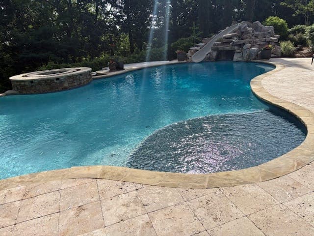 Freeform gunite pool with a travertine paver deck, a raised stacked stone spa, and a rock waterfall in a wooded backyard