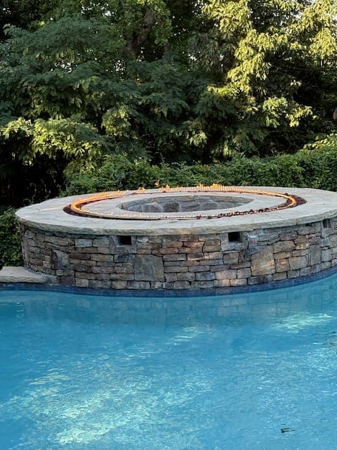 Round stacked stone spa with a gas fire feature set into the top, beside a freeform pool