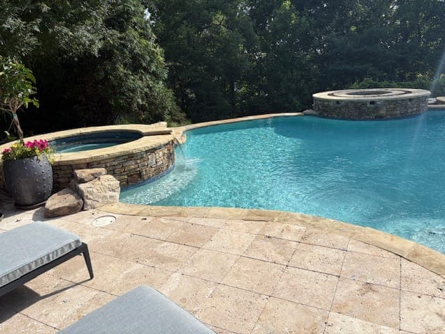 Raised stacked stone spa spilling into a freeform pool, with a travertine paver deck and lounge chairs