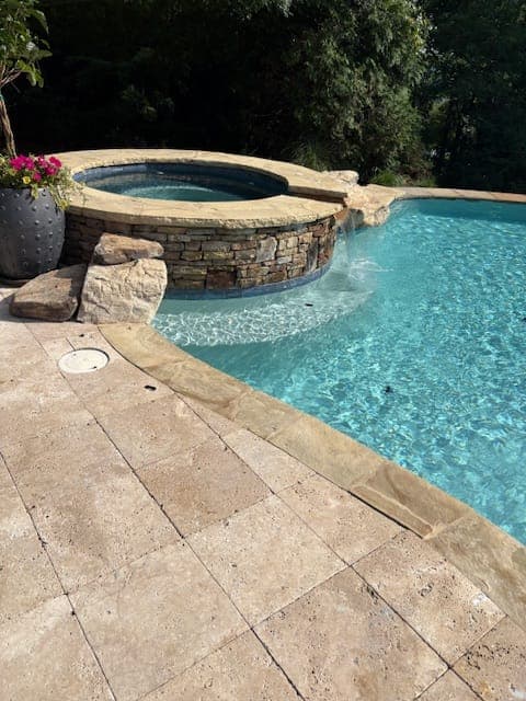 Water spilling from a raised stacked stone spa into a freeform pool edged with travertine pavers